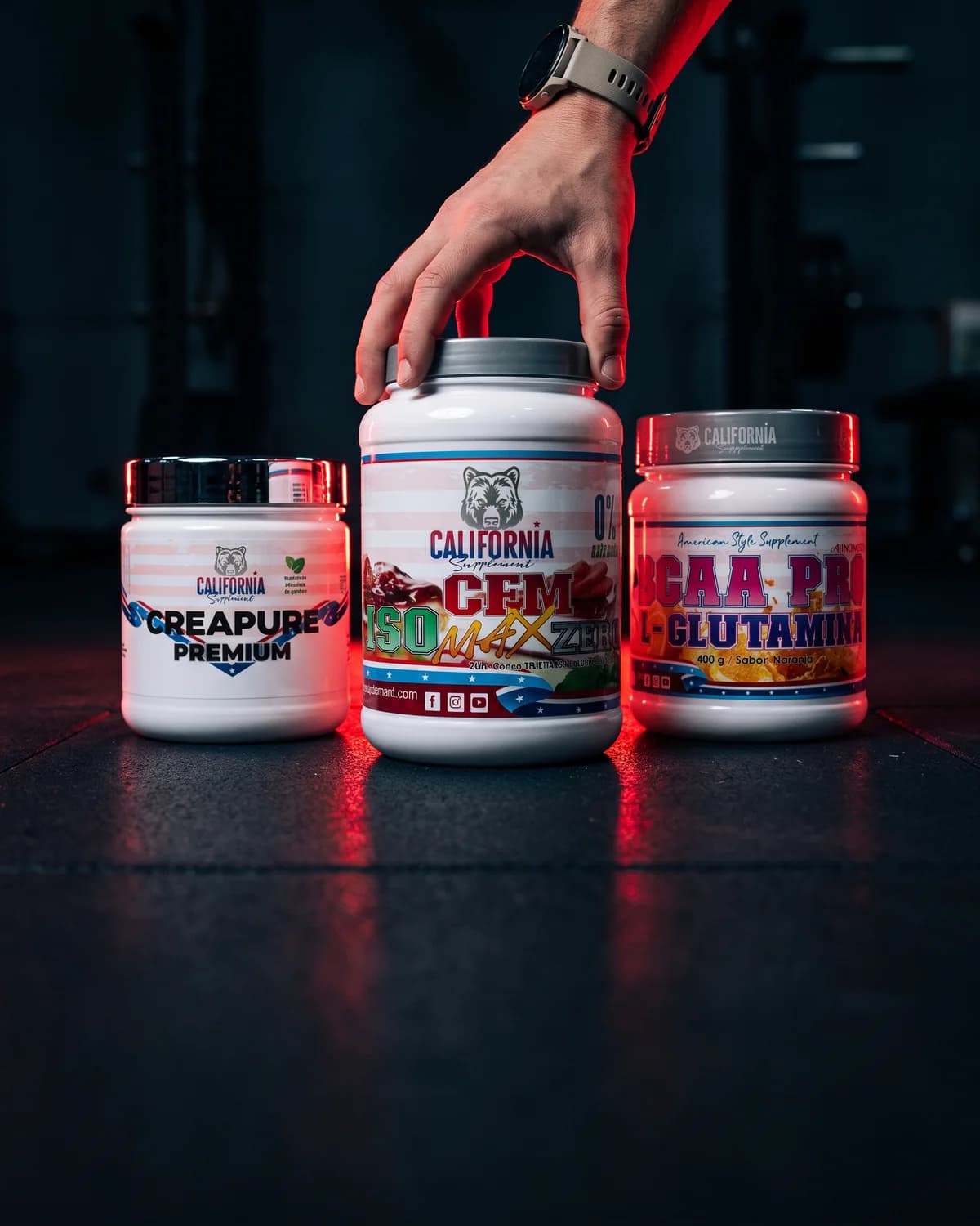 Tres botes de California Supplement, Creapure Premium, CFM Iso Max Zero y BCAA Pro con L-Glutamina, y una mano levantando la tapa del bote central